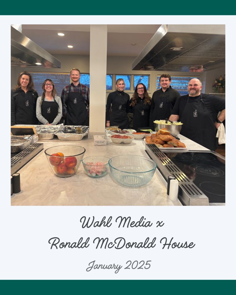 Seven chefs in aprons stand behind a food-filled kitchen island. Caption: “Wahl Media x Ronald McDonald House, Jan 2025—Fairport, NY, experts in strategic media buying and brand growth.”.