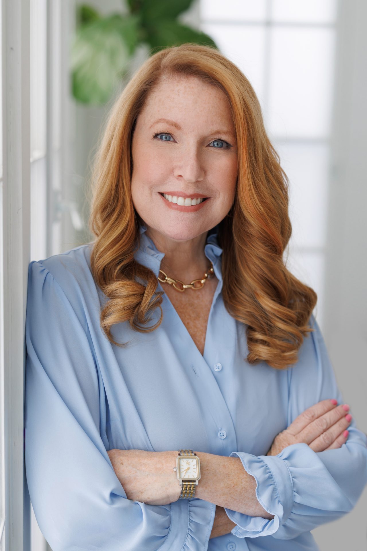 A smiling woman with red hair and blue eyes in a blue blouse leans on a white wall, representing Fairport’s strategic media experts.