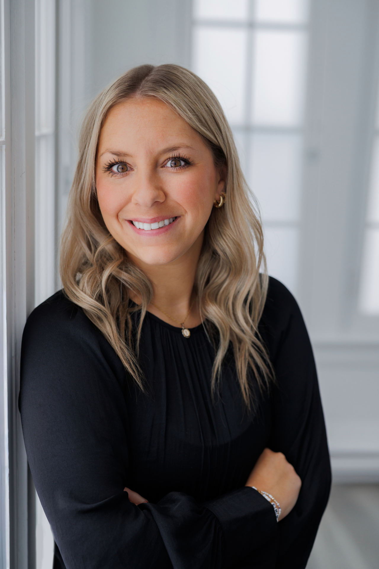 A confident, smiling woman with long blonde hair in a black top stands arms crossed in a bright, windowed room, embodying expertise.